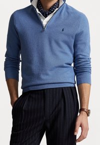 Ljusblå tröja med dragkedja vid kragen, stickad textur och ribbade ärmslut, i kombination med mörka randiga byxor och en prickig scarf.