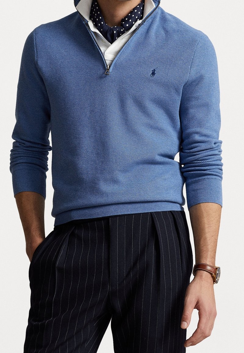 Ljusblå tröja med dragkedja vid kragen, stickad textur och ribbade ärmslut, i kombination med mörka randiga byxor och en prickig scarf.