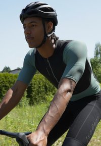 Kortärmad cykeltröja i grön och svart, med dragkedja framtill, texturerat tyg och figursydd design. Bärs med en svart hjälm.