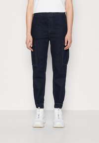 Mörkblå cargobyxor med elastisk midja, två sidofickor, avsmalnande muddar och textur av denim. Bärs med vita sneakers.