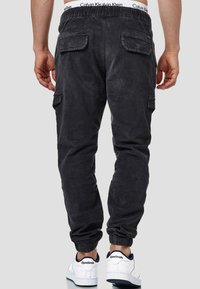 Pantalon cargo gris foncé avec une taille élastique, deux poches arrière, des poignets de jambe fuselés et des poches latérales. Porté avec des sneakers blanches Reebok.