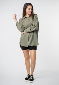 Olijfgroen overhemd met knopen in een gestructureerde stof, gecombineerd met zwarte shorts en zwarte sneakers. Bril in de hand, casual houding.