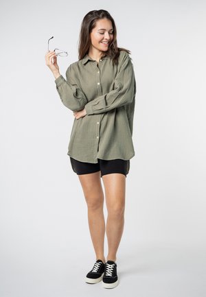 Olive green button-up shirt in textured fabric, paired with black shorts and black sneakers. Glasses held in hand, casual pose.