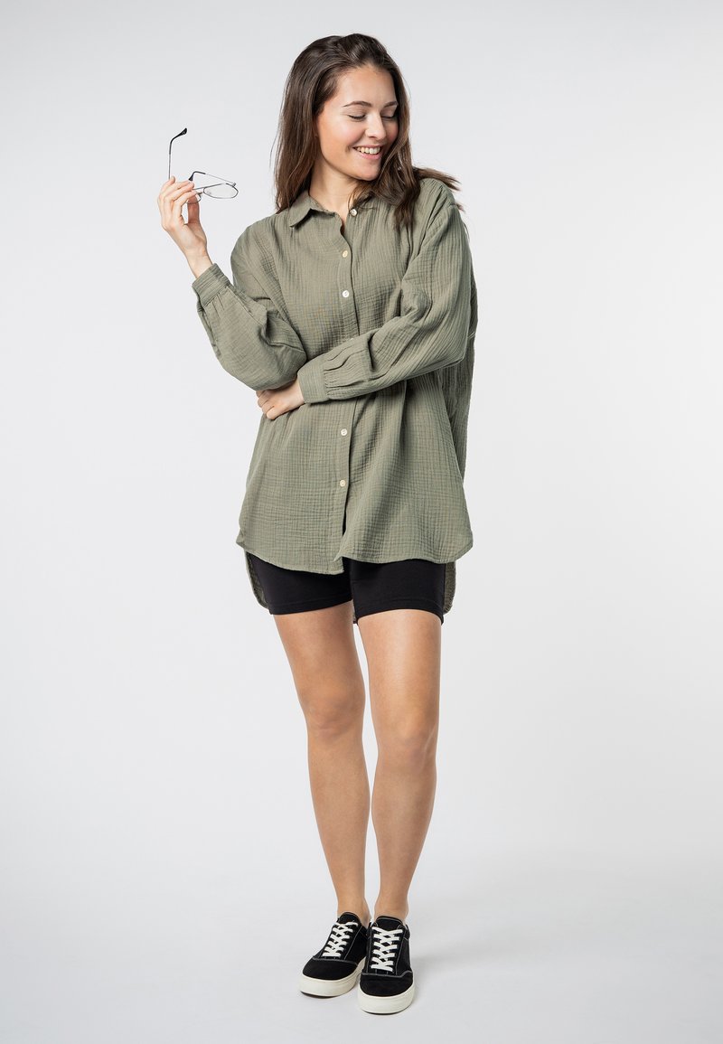 Olijfgroen overhemd met knopen in een gestructureerde stof, gecombineerd met zwarte shorts en zwarte sneakers. Bril in de hand, casual houding.