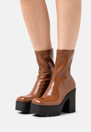 Braune, glänzende Stiefeletten mit schlankem Design, einer dicken schwarzen Plateausohle und hohem Blockabsatz, gefertigt aus glattem, glänzendem Material.
