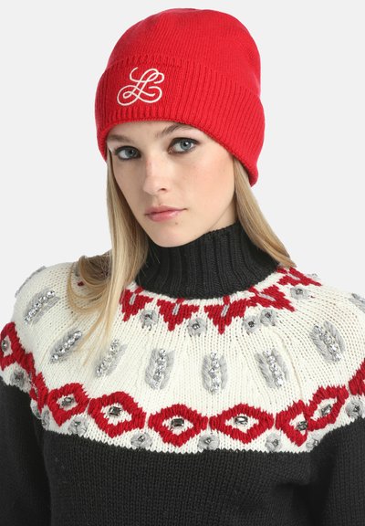 Rdeča pletena kapa z vezenim belim logotipom "L". Nosi se z črnim puloverjem z visokim ovratnikom, ki ima smetanasto in rdečo vzorčeno zasnovo ter srebrne detajle.