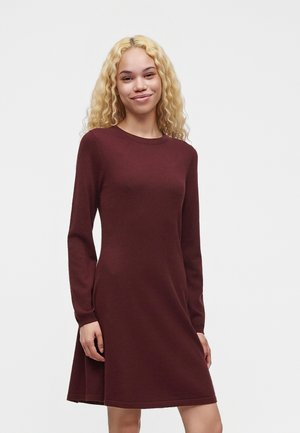 Jonge vrouw met lang krullend blond haar, een donkerrode jurk met lange mouwen en knie-lang, staand tegen een effen achtergrond.