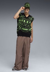 Top sin mangas de punto negro con un diseño gráfico en verde brillante, combinado con pantalones cargo sueltos en beige y un gorro a juego.