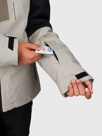 Una mano che inserisce una carta in una piccola tasca di una giacca beige, con chiusura in velcro nero, contro uno sfondo semplice.