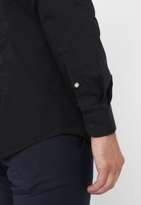 Zwarte langärmige shirt met knoopmanchetten, vervaardigd uit een gladde stof, met een getailleerde pasvorm en een subtiele glans. De knopen zijn rond en contrasterend.