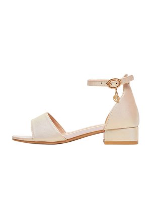 Beige sandal med lav hæl, åben tå, ankrem med guldlås og lille guldsmykke, der hænger fra remmen.