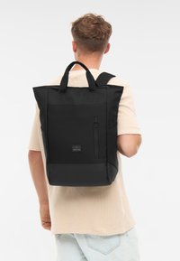 Fekete hátizsák téglalap alakú, vászon külső résszel, elülső cipzáras zsebbel és dupla felső fogantyúval. Jellemzője egy texturált alj és logós díszítés.