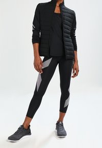 Svart idrottsjacka med quiltad textur, i kombination med en åtsittande svart topp och svarta leggings med silver geometriska accenter och grå löparskor.