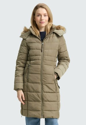 Frau in einer olivgrünen, gesteppten Winterjacke mit einem kapuzenbesatz aus Fell, die mit einer Hand in der Tasche vor einem schlichten Hintergrund steht.