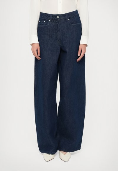 Kvinde iført mørkeblå wide-leg jeans, hvid skjorte med knapper og hvide spidse sko, stående foran en ensfarvet baggrund.