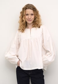 Balta medvilninė bluzė su apvaliu iškirpimu, klokštelėmis prie kaklo ir apimtinėmis rankovėmis. Pora su tamsiomis džinsinio audinio kelnėmis.