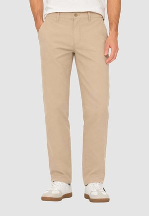 Uomo che indossa pantaloni beige a gamba dritta e sneakers bianche, con le mani nelle tasche, in piedi contro uno sfondo chiaro e uniforme.