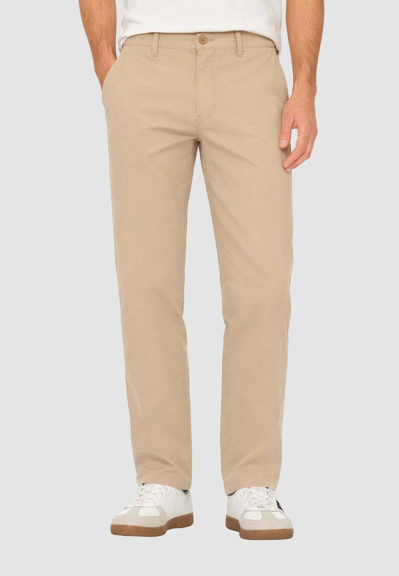 Homme portant un pantalon beige droit et des baskets blanches, les mains dans les poches, debout devant un fond clair uni.