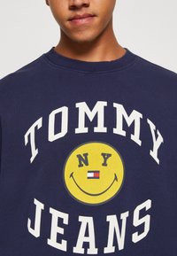 Námořnická modrá mikina vyrobená z bavlny, s bílým textem "TOMMY" a "JEANS" a žlutým úsměvem uprostřed se "NY".