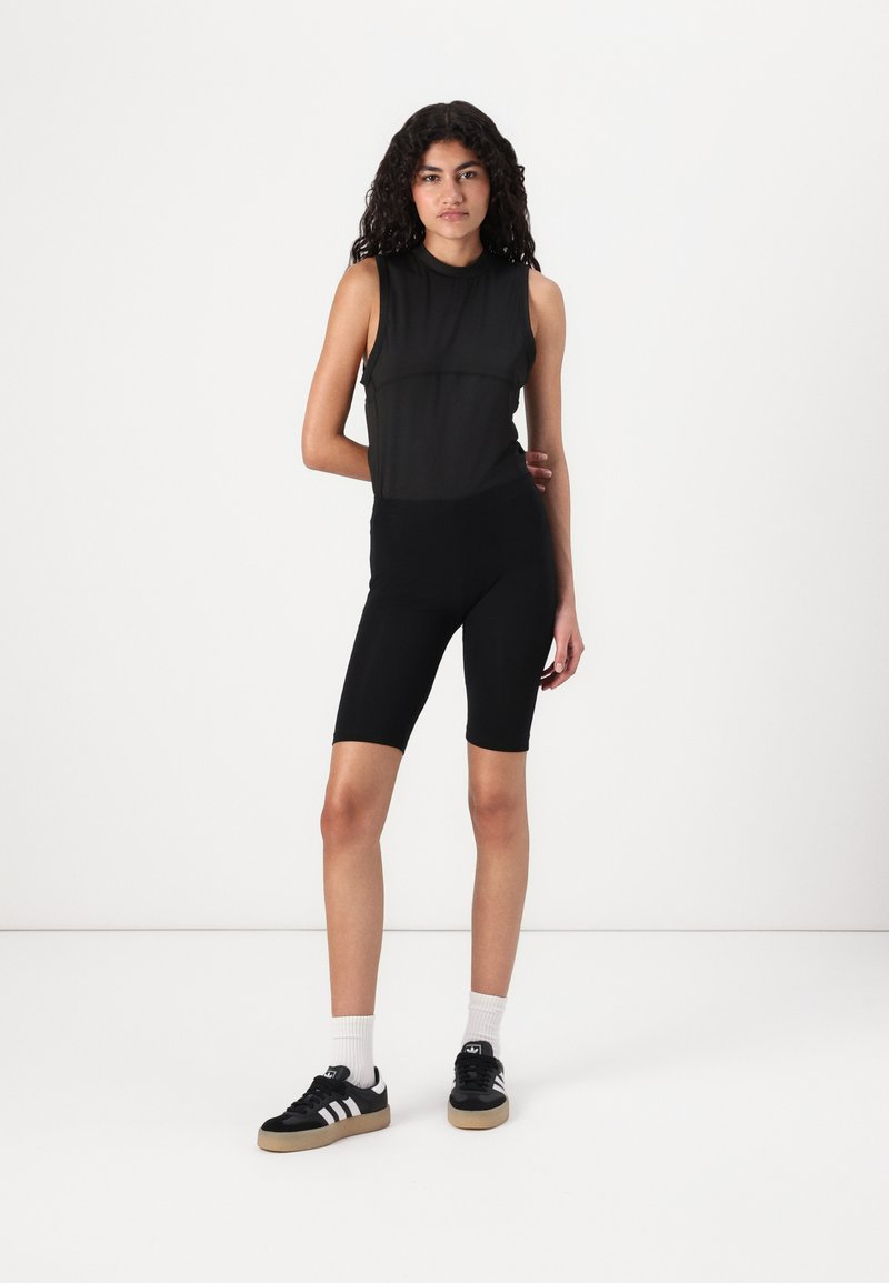 Svart atletisk bodysuit med ermeløs design, kombinert med knelengde sorte sykkelshorts og svarte sneakers med hvite detaljer og striper.