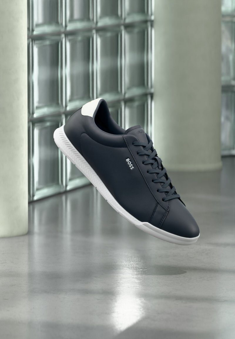 Baskets en cuir noir avec semelle blanche et languette au talon, logo de la marque « BOSS » sur le côté, présentées flottant devant un fond de blocs de verre et de béton.