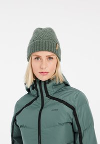 Gorro de punto verde con un patrón texturizado y una etiqueta de cuero marrón claro. El modelo lleva una chaqueta acolchada verde azulado con acentos negros.