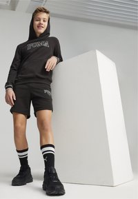 Sudadera negra con logo, pantalones cortos negros a juego y zapatillas negras combinadas con calcetines a rayas blancas y negras. Diseño sencillo y estilo urbano.