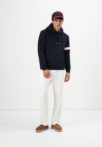 Mann iført mørk marineblå hettegenser med rød og hvit armstripe, hvite bukser, brune slip-on sko og en burgunder baseballcap, står foran en hvit bakgrunn.