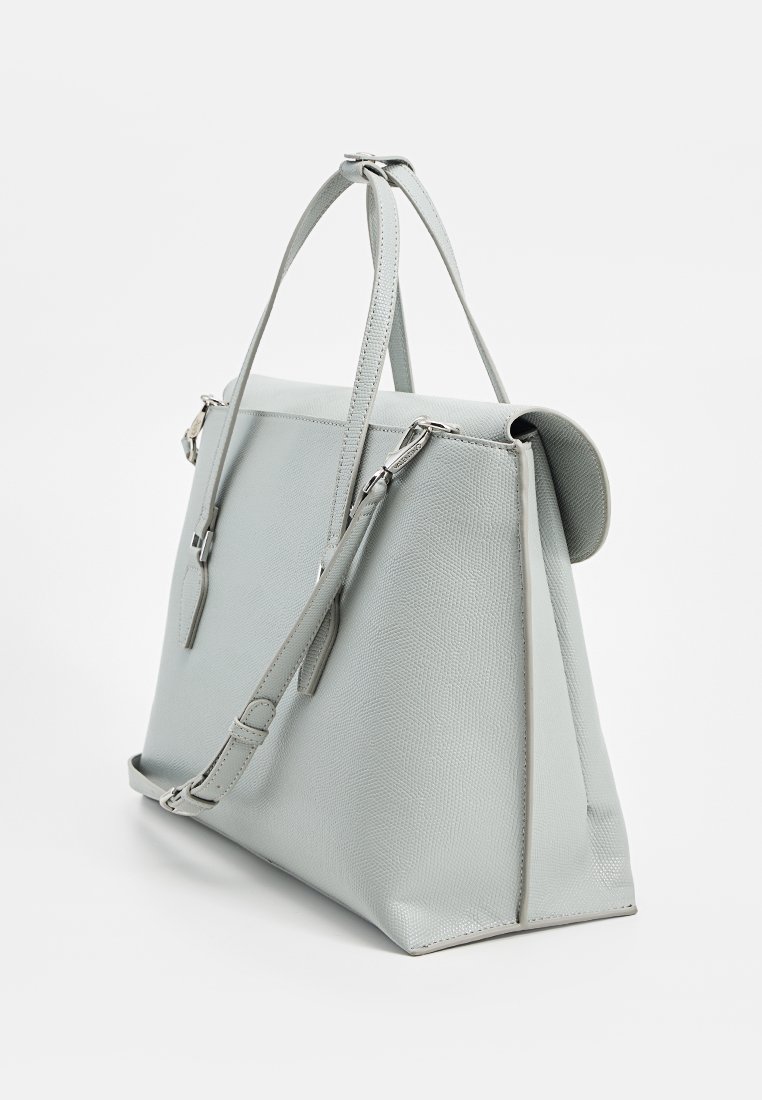 Sac à main en cuir texturé gris clair avec deux poignées, une bandoulière réglable et une forme trapézoïdale structurée sur fond blanc.