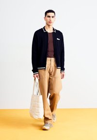 Czarna kurtka na zamek z białymi wykończeniami, brązowa koszulka z prążkowanej tkaniny, beżowe spodnie o szerokich nogawkach oraz kremowa pikowana torba. Jasnoszare sneakersy.