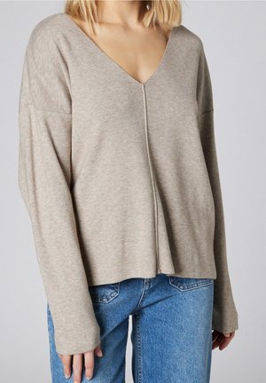 Beiger V-Ausschnitt-Pullover mit lockerer Passform, langen Ärmeln und einer Nahtdetails in der Mitte, kombiniert mit hochgeschnittenen blauen Jeans.