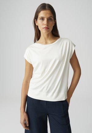 Giovane donna con lunghi capelli castani, indossa una maglietta bianca a maniche corte e pantaloni blu navy, in piedi davanti a uno sfondo grigio uniforme.