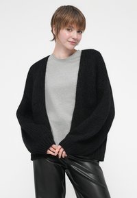 Fekete, vastag kötésű kardigán, oversized ujakkal és nyitott elülső résszel, szürke pólóra rétegezve, fekete bőrnadrággal párosítva.