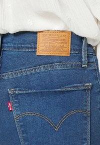 Los jeans de mezclilla azules presentan un parche de cuero marrón con texto en relieve, un bolsillo trasero con una etiqueta roja y costuras en contraste en las costuras.