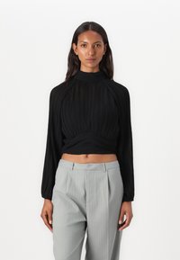 Top nero a maniche lunghe con texture plissè, colletto alto e vita arricciata abbinato a pantaloni grigi chiari con righe sottili. Design semplice e forma aderente.