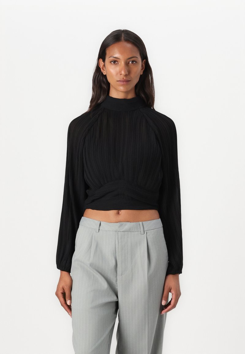 Top nero a maniche lunghe con texture plissè, colletto alto e vita arricciata abbinato a pantaloni grigi chiari con righe sottili. Design semplice e forma aderente.