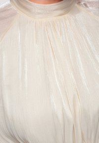 Tissu crème scintillant avec des plis froncés autour d'un col montant, mettant en valeur une texture douce et un éclat subtil.