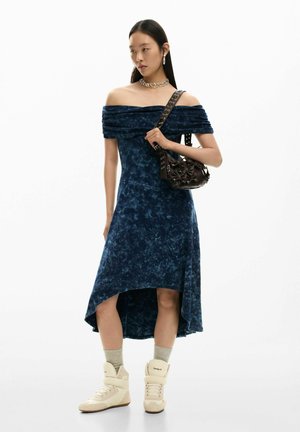 Tmavomodré šaty s odhalenými ramenami, so zloženým výstrihom, asymetrickým lemom a jemnou textúrou, doplnené béžovými vysokými topánkami a tmavou kabelkou.