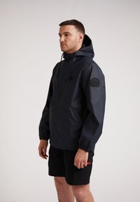 Veste imperméable navy avec une capuche ajustée, des poignets élastiques et une patch logo sur la manche. Portée sur un short noir, maintenu avec un objet rouge.