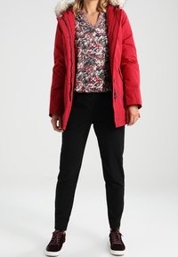 Mujer vestida con una chaqueta de invierno con capucha roja, blusa floral, pantalones negros y zapatillas oscuras, de pie con las manos relajadas a los lados.