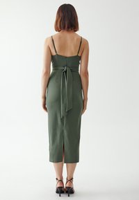 Olivengrünes, figurbetontes Kleid mit dünnen Trägern, einer gebundenen Taillendetail und einem Schlitz im Rücken. Weicher Stoff mit minimalistischem Design.