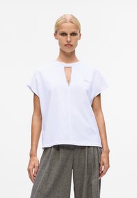 Weißes Baumwoll-T-Shirt mit kurzen Ärmeln, ausgestattet mit einem Schlüsselloch-Ausschnitt und silbernen Akzenten. Kombiniert mit grauen, strukturierten Schlaghosen.