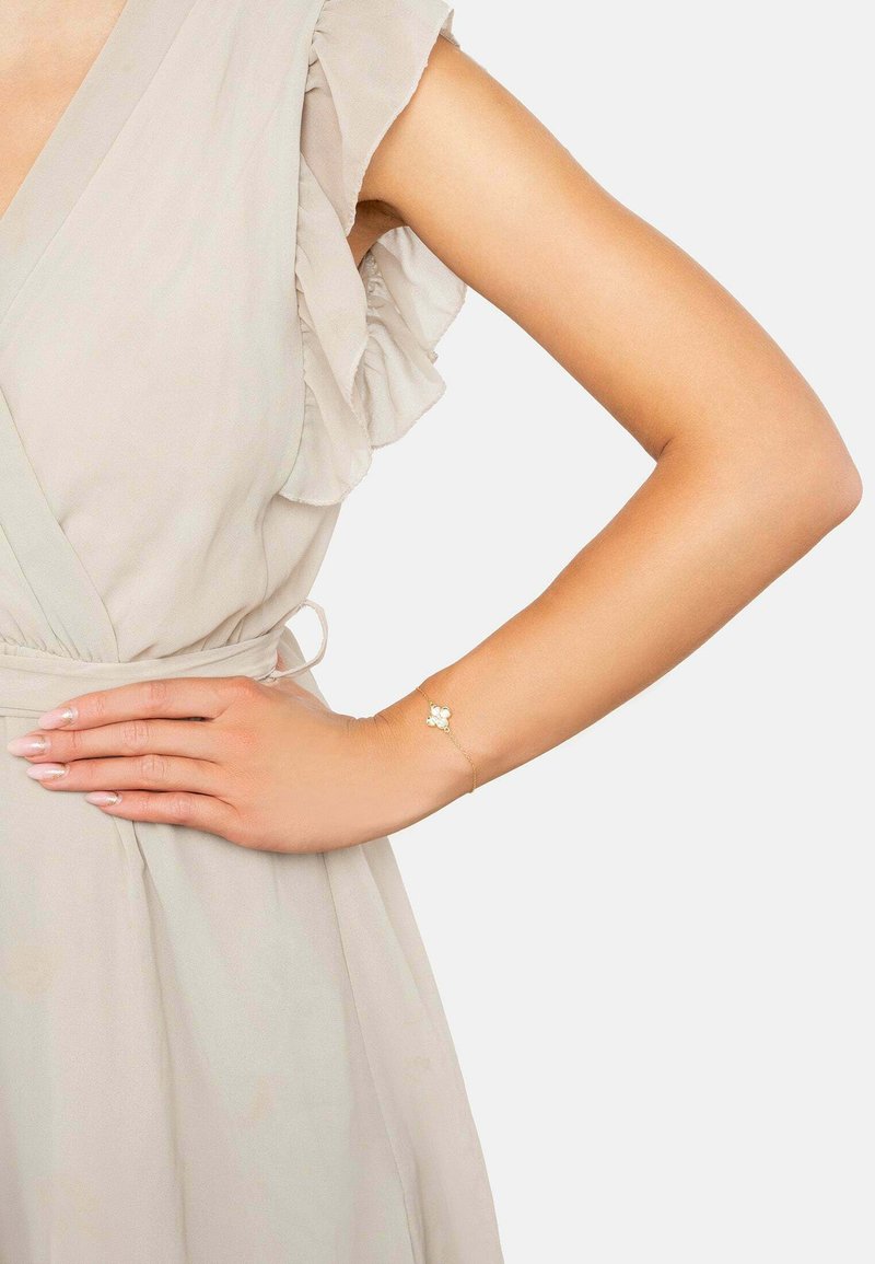 Gold bracelet with floral accent, featuring white embellishments. Worn on a wrist against a light beige dress with ruffled sleeves.