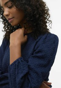 Abbigliamento blu navy con maniche lunghe a sbuffo, realizzato in un tessuto stropicciato. Il modello ha i capelli ricci e sta aggiustando delicatamente lo scollo.