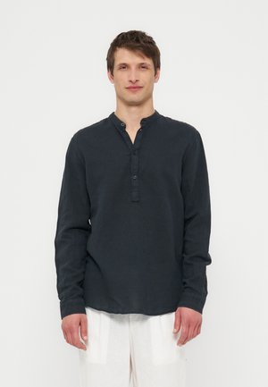 Mladý muž s krátkymi hnedými vlasmi v čiernom henley tričku s dlhým rukávom a bielych nohaviciach stojaci na bielom jednoduchom pozadí.