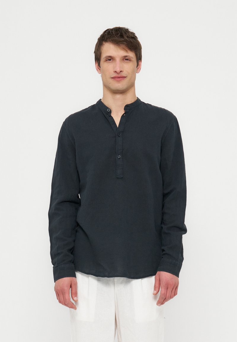 Νέος άνδρας με κοντά καστανά μαλλιά που φοράει μαύρο μακρυμάνικο μπλουζάκι Henley και λευκό παντελόνι, στέκεται σε λευκό φόντο.