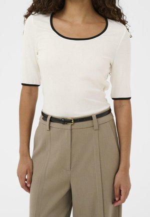 Femme portant un haut blanc à manches courtes avec bordure noire et un pantalon beige taille haute avec une ceinture noire.
