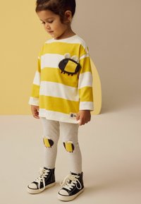 Gestreiftes, langärmliges Shirt in Gelb und Weiß mit einer schwarzen und gelben Tasche. Graue Leggings mit Bienen an den Knien. Schwarze High-Top-Sneaker.