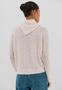 Maglione lavorato a maglia in beige chiaro con un design a trama larga, dotato di cappuccio e orlo a coste. Abbinato a jeans di denim, che mostrano una vestibilità rilassata.