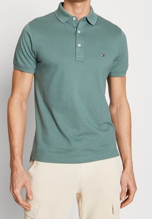 Homme portant un polo à manches courtes vert sourd avec trois boutons et un pantalon cargo beige, debout devant un fond clair uni.